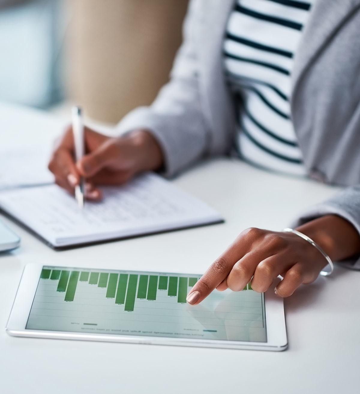 A person analyzes financial data on a tablet with a bar graph, while taking notes in a notebook, to make money work for her
