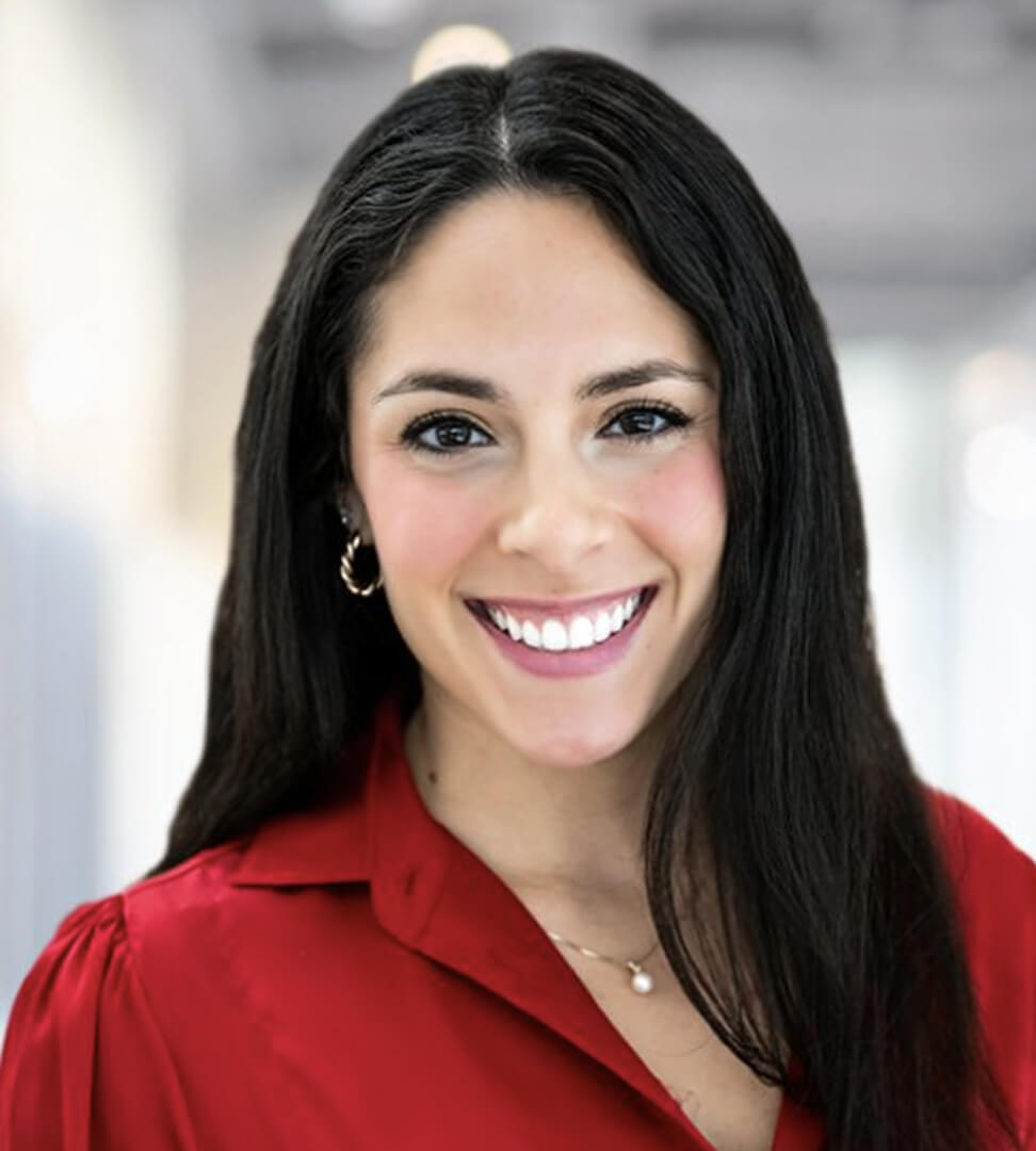A woman with long dark hair, wearing a red blouse and jewelry, smiling brightly in an indoor setting.