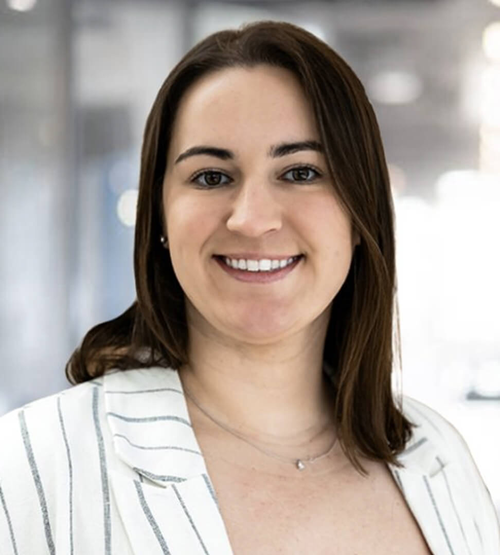 A woman with shoulder-length brown hair, smiling, wearing a white blazer with black stripes, in a professional setting.