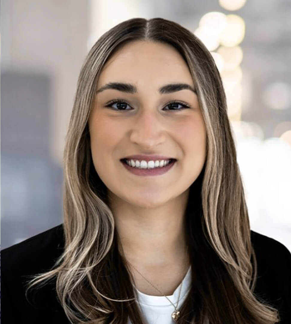 A smiling young woman with long, wavy brown hair, wearing a black blazer and white top, in a professional setting.