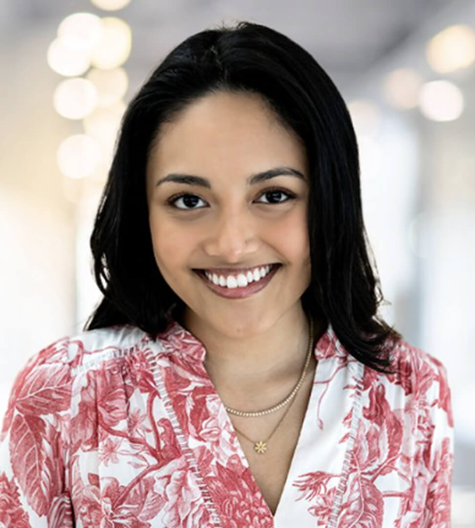 A smiling woman with shoulder-length black hair wearing a red and white floral blouse and layered necklaces.