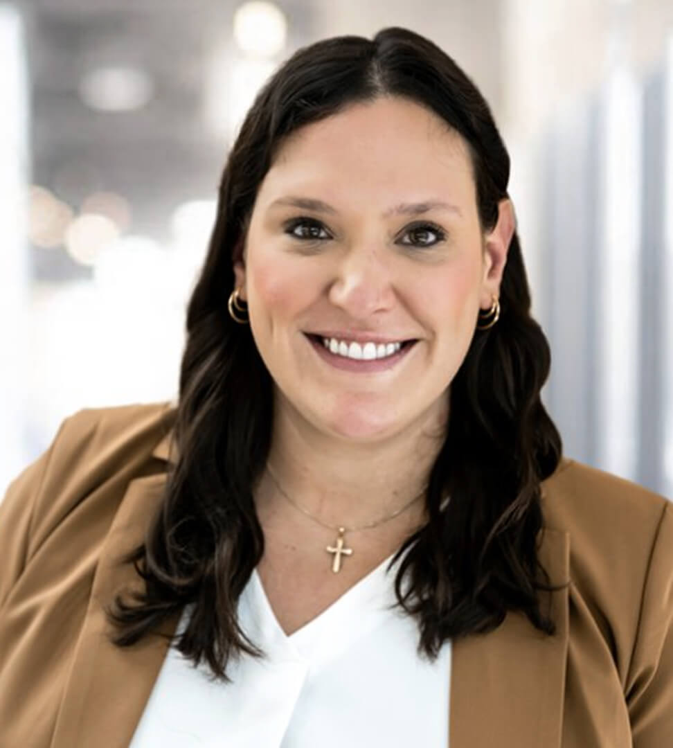 A woman with dark wavy hair, wearing a cross necklace, gold hoop earrings, and a tan blazer, smiling in an indoor setting.