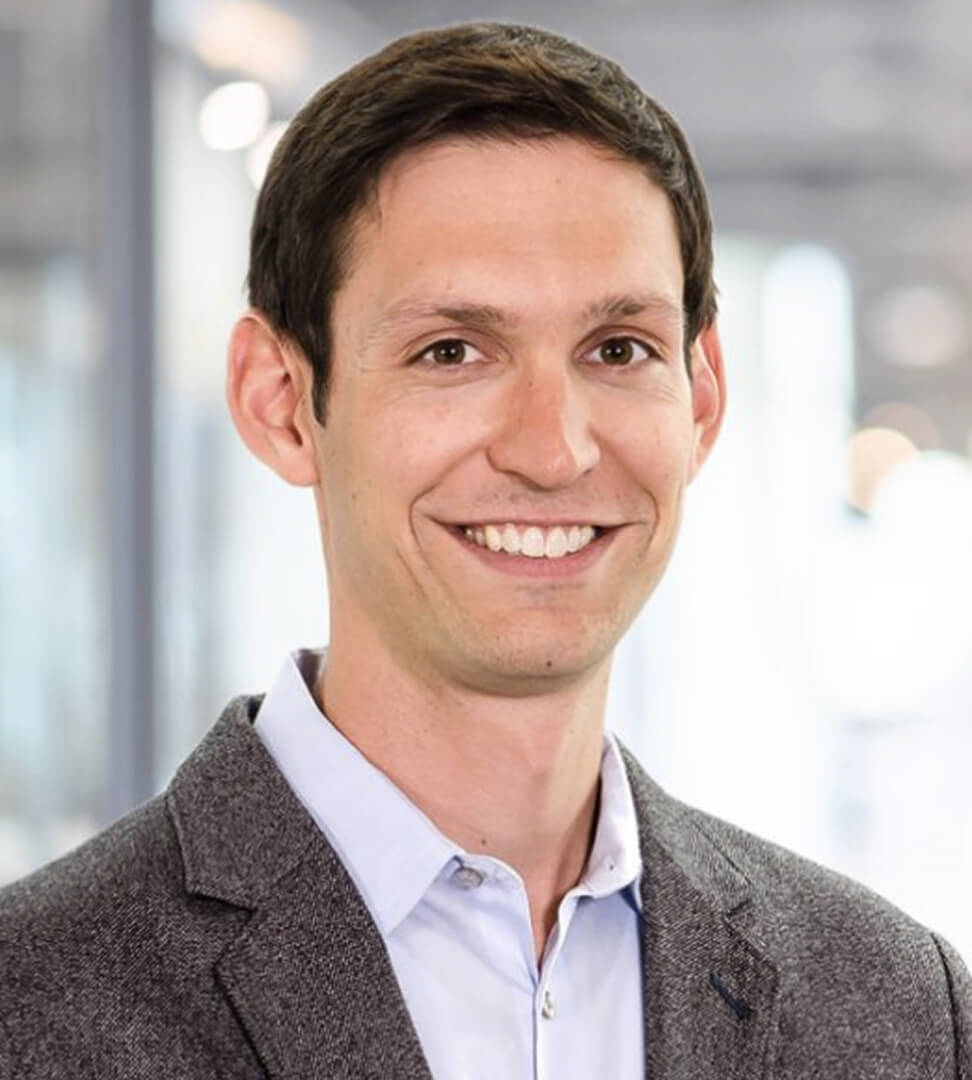 A smiling man in a gray blazer and white shirt in a professional setting with blurred background.
