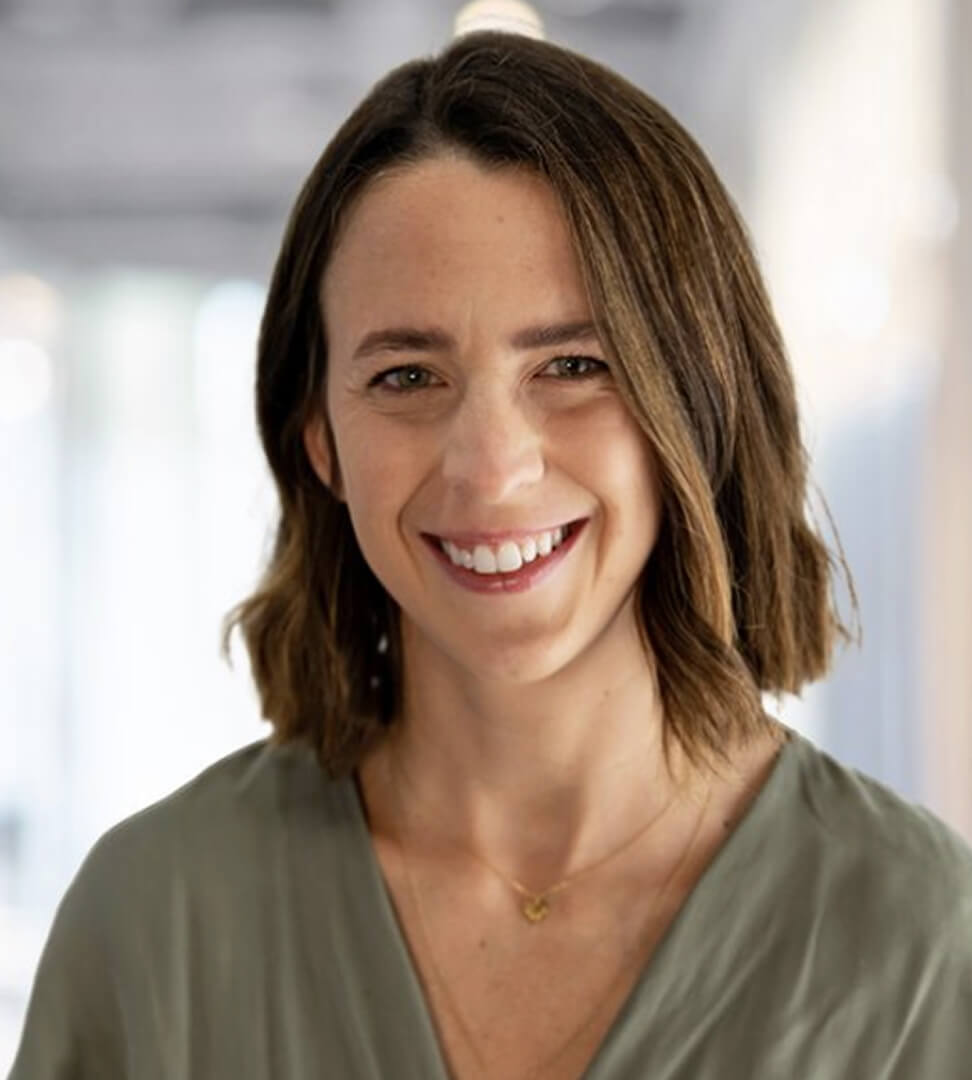 A woman with shoulder-length brown hair smiling in a professional setting, wearing a gray top and a delicate gold necklace.