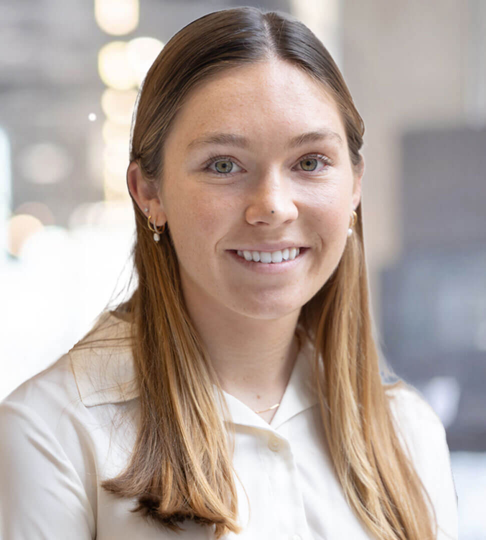 A young woman with long red hair, wearing a white shirt and gold earrings, smiling in a bright indoor setting.