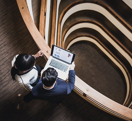 Synapse Why Choose Us Two professionals review data on a laptop in a modern, curved architectural space, highlighting collaboration and innovation.